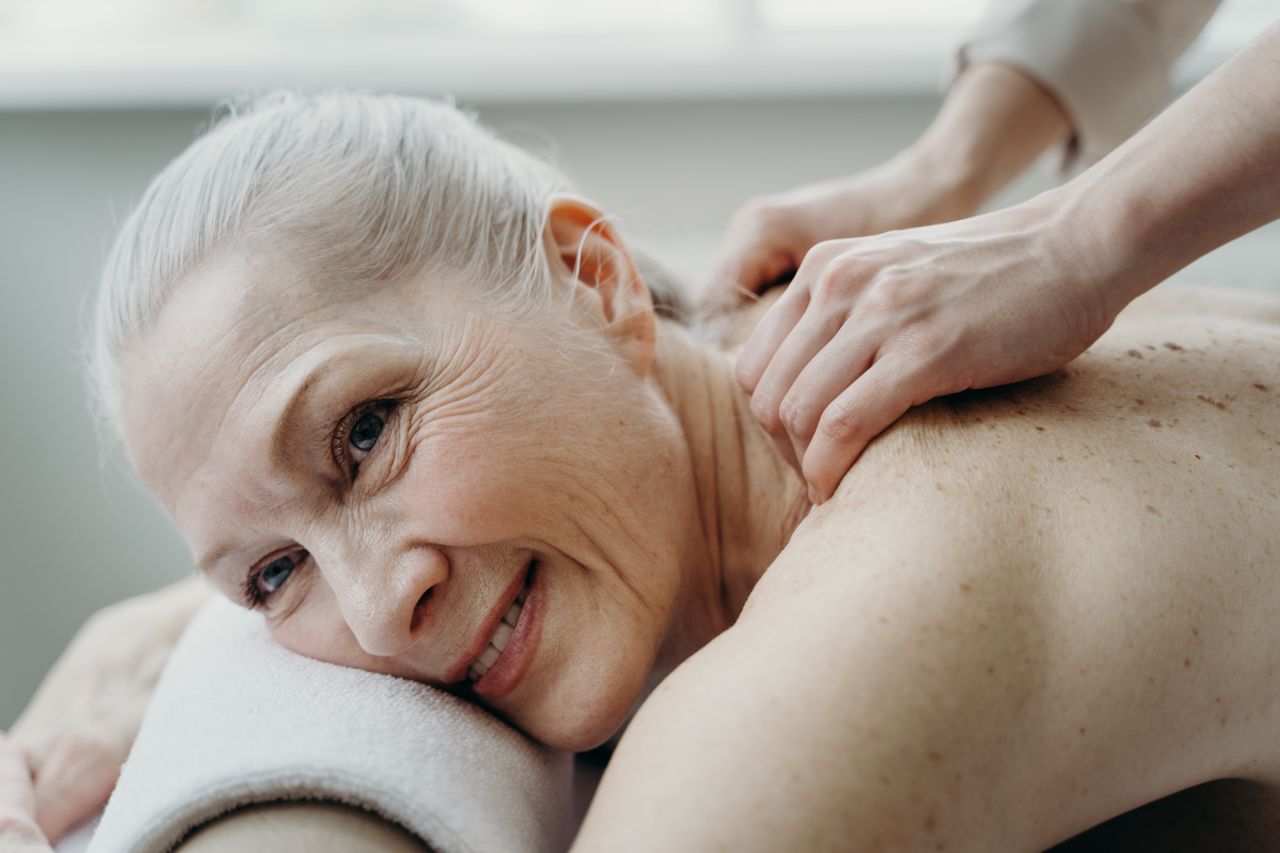 contact-img A senior woman with white hair smiles while receiving a therapeutic back massage indoors.